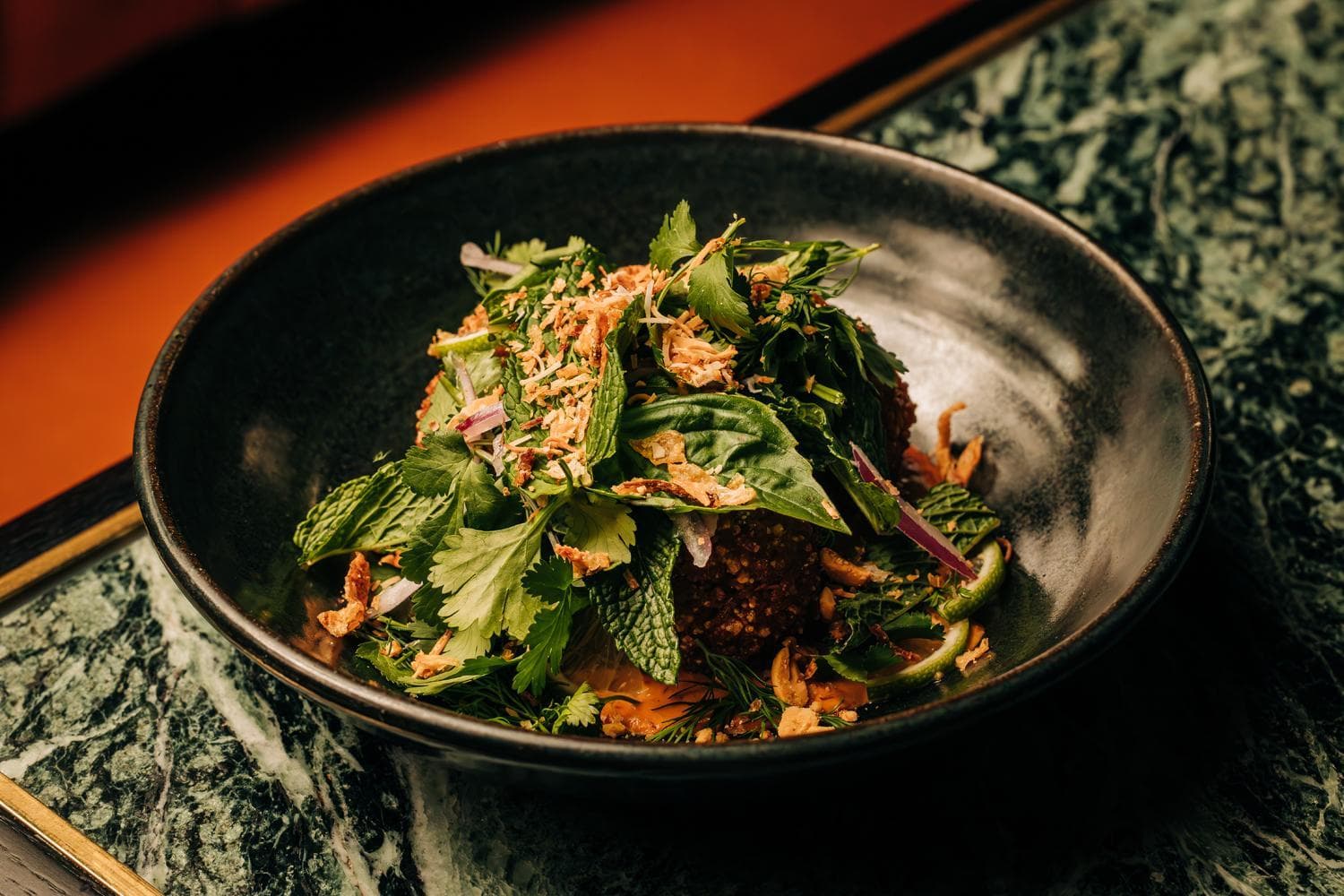 A dark ceramic bowl contains a crispy fried rice dish garnished with fresh green herbs and sliced red onions. The surface underneath appears to be mottled green marble.
