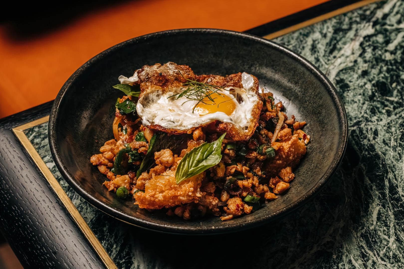 A black bowl filled with a savory dish, including crispy rice, green herbs, and vegetables, topped with a fried egg.
