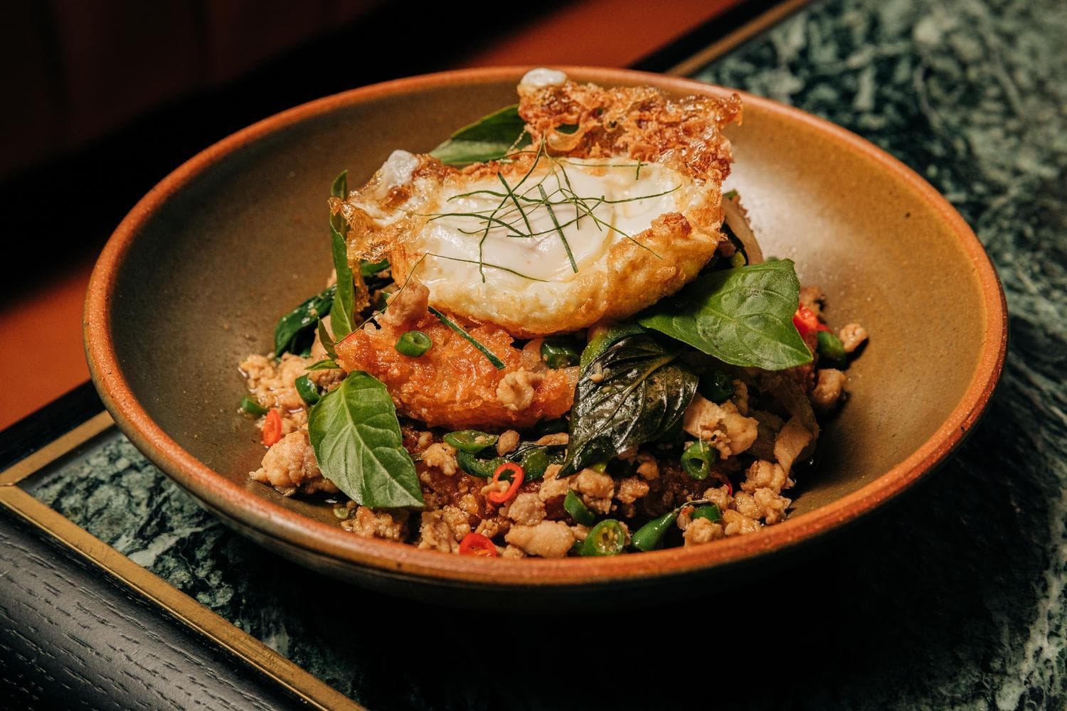 A black bowl filled with a savory dish, including crispy rice, green herbs, and vegetables, topped with a fried egg.