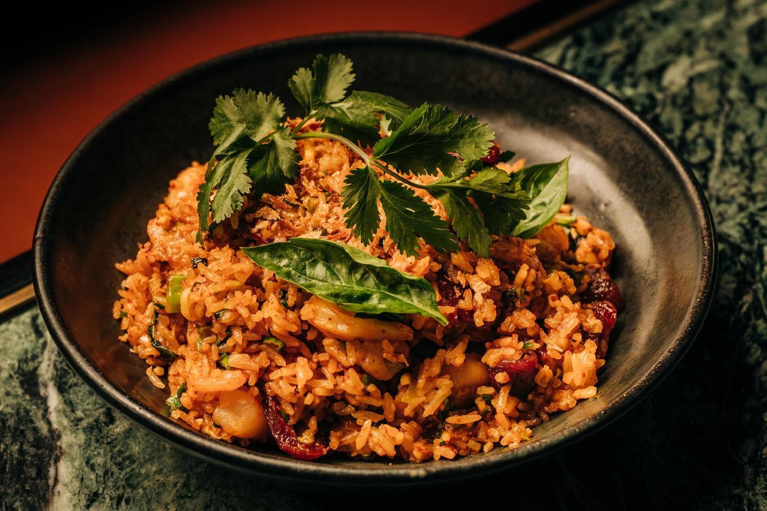 A bowl of fried rice garnished with cilantro leaves, featuring fresh herbs, in a dark-colored dish.
