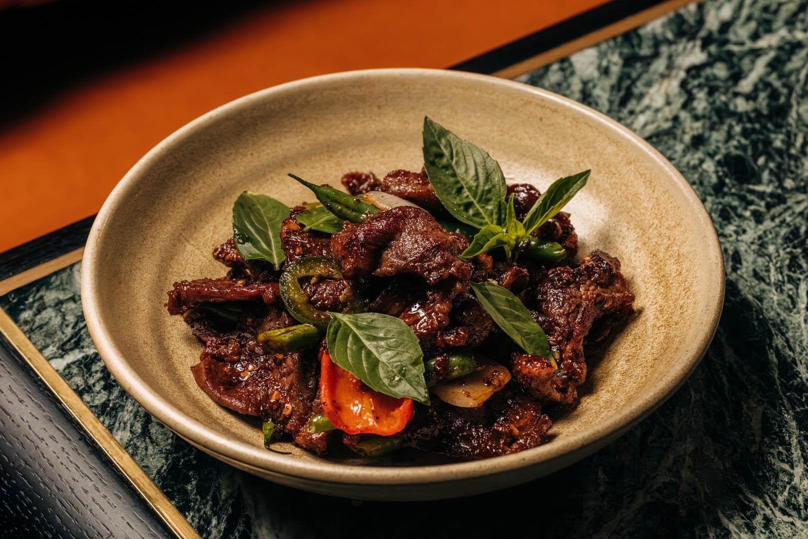 A bowl of cumin lamb with basil leaves, sliced chili peppers, and herbs, placed on a patterned surface. The dish is vibrant with a mix of red and green colors.